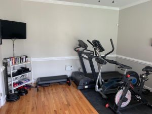 View of workout side of room with exercise equipment, wall-mounted tv, dvds and weights on shelving.